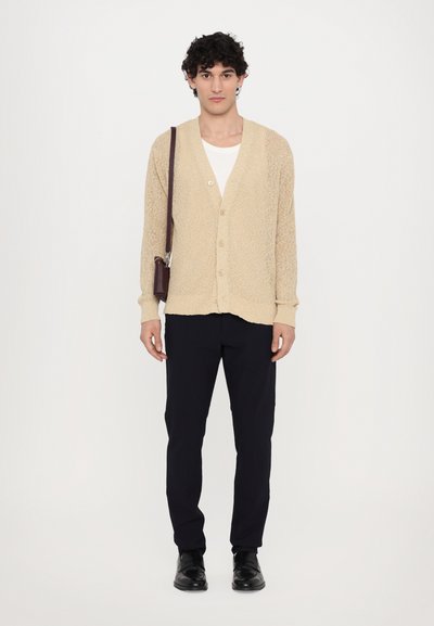 Lys beige strikket cardigan med V-hals og fire knapper, kombinert med svarte bukser og svarte sko, holdt med en brun skulderveske.