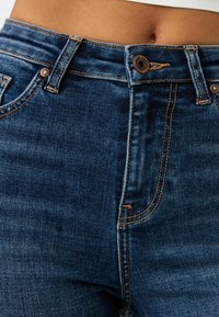 Mörkblå denimjeans med strukturerad midja, kontrasterande orange sömmar, silverknappstängning och standard femfickdesign.