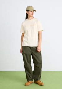 Krämfärgad t-shirt av bomull med relieferat "Timberland"-logotyp, kombinerad med olivgröna cargobyxor, tan-färgade snörskor och en ljusgrön keps.