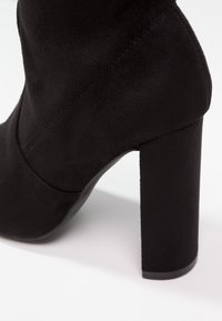 Schwarz Wildleder High-Heel-Stiefel mit dicker Absätze, glatter Oberfläche und Nahtdetails im oberen Bereich.