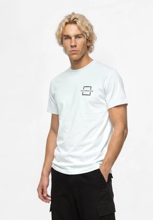 Jeune homme aux cheveux blonds bouclés portant un t-shirt blanc à manches courtes avec un petit logo noir et un pantalon noir, debout devant un fond uni.