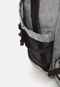 Grijze stoffen rugzak met een ruitpatroon, voorzien van een zijvak van gaas en zwarte banden voor een verstelbare pasvorm. Lichtgewicht en duurzaam ontwerp.