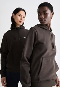 Sudaderas con capucha marrones hechas de tejido suave con cordones, bolsillos tipo canguro y pequeño logo verde en el pecho.