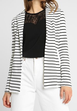 Femme portant un blazer blanc à rayures horizontales noires, un haut noir orné de dentelle et un pantalon blanc taille haute, debout devant un fond uni.