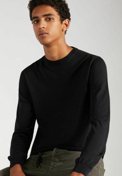Chemise noire à manches longues et col rond avec une texture lisse. Le modèle porte un pantalon vert olive. Design simple, sans motifs ni accents visibles.