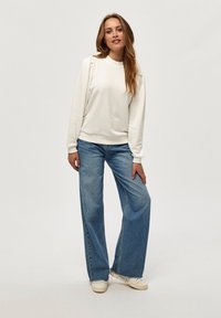 Sudadera blanca con sutiles detalles de volantes en los hombros, combinada con jeans de denim de pierna ancha en azul claro, y finalizada con zapatillas blancas.