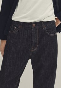 Tummansiniset farkut, joissa on viisi taskua, oranssitomitus ja metallinen nappi. Kangas on tekstuuripintaista ja hieman kulunutta.