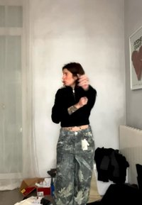 Woman adjusting her sleeve wearing black top and gray camo pants with tattoos visible, standing in a cluttered room with a framed heart poster.