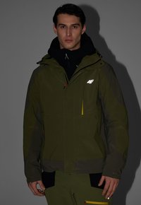 Homme portant une veste et un pantalon d'extérieur vert foncé avec des détails noirs et jaunes sur fond gris.