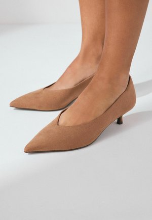 Tacones puntiagudos en ante color arena con un bajo tacón de kitten, que presentan un corte en V profundo a lo largo del borde superior. Textura suave sin patrones visibles.