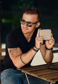 Bežová kožená peněženka s hladkým povrchem, kterou drží osoba v černém tričku, sedící u dřevěného stolu.