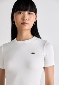 Aasia naine sirgete mustade juustega, kes kannab väikeseid rõngastikke ja valget ribakestega lühikeste varrukatega särke, millel on roheline krokodilli logo rinnal.