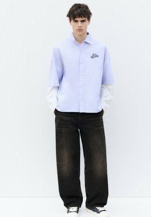 Jeune homme aux cheveux courts et foncés portant une chemise bleu clair à manches courtes sur des manches longues blanches, un jean noir large et des baskets noires, debout contre un mur blanc.