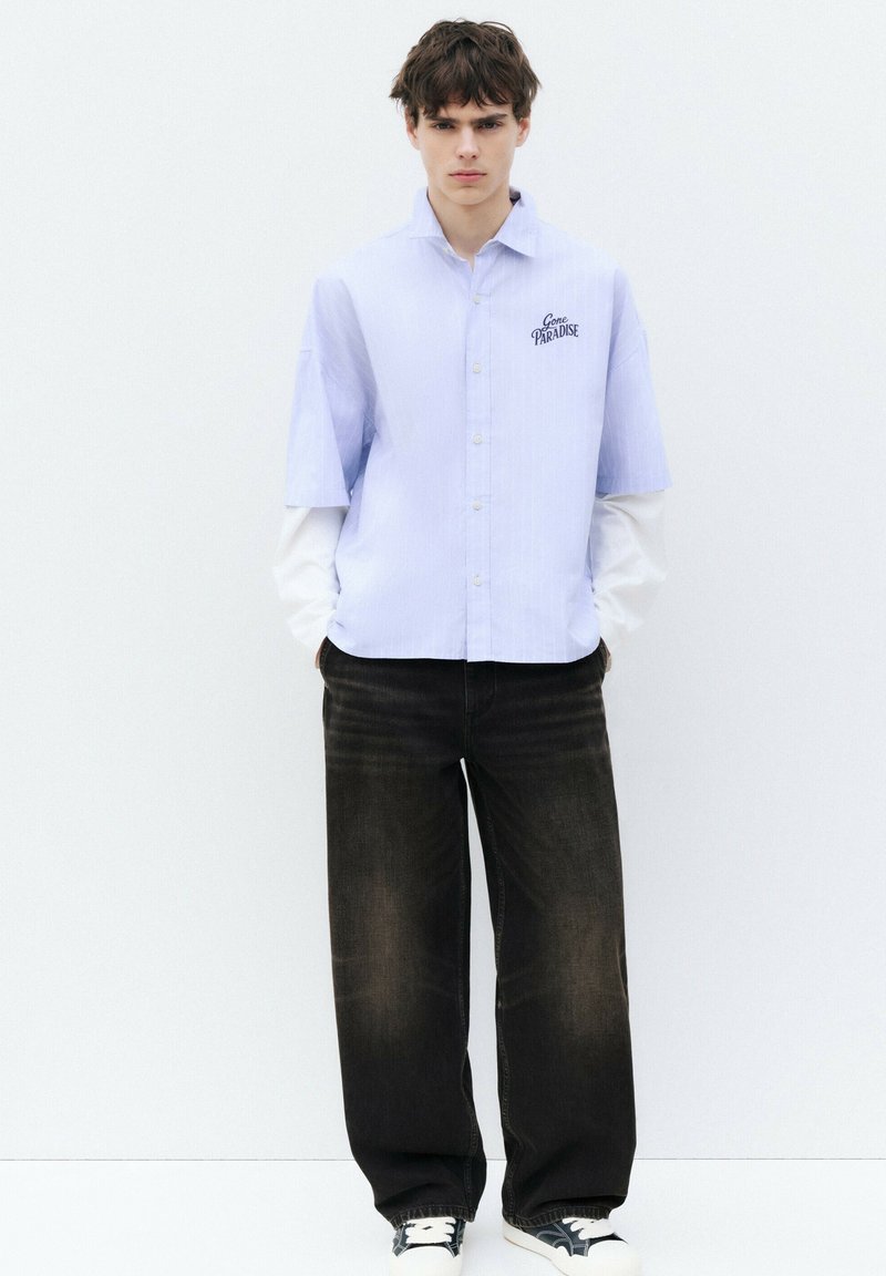 Jeune homme aux cheveux courts et foncés portant une chemise bleu clair à manches courtes sur des manches longues blanches, un jean noir large et des baskets noires, debout contre un mur blanc.