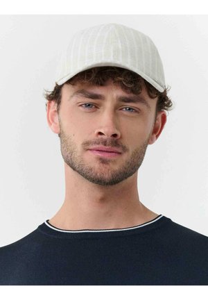 Mladý muž s jemným fúzom, ktorý má na hlave bielu čiapku a čierne tričko bez goliera, stojaci pred jednoduchým svetlým pozadím.