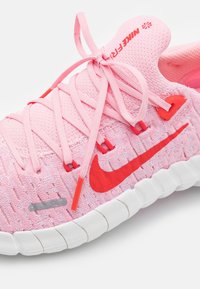 Chaussure de sport rose avec une tige en tissu texturé, présentant des accents rouges et une semelle blanche segmentée. Les lacets sont soigneusement noués en doubles nœuds.