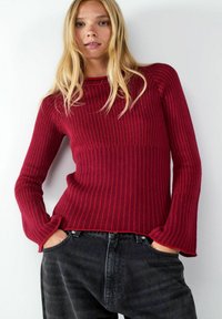 Roter gerippter Strickpullover mit rundem Halsausschnitt und langen Ärmeln, kombiniert mit hochtaillierten schwarzen Jeans in lockerer Passform.