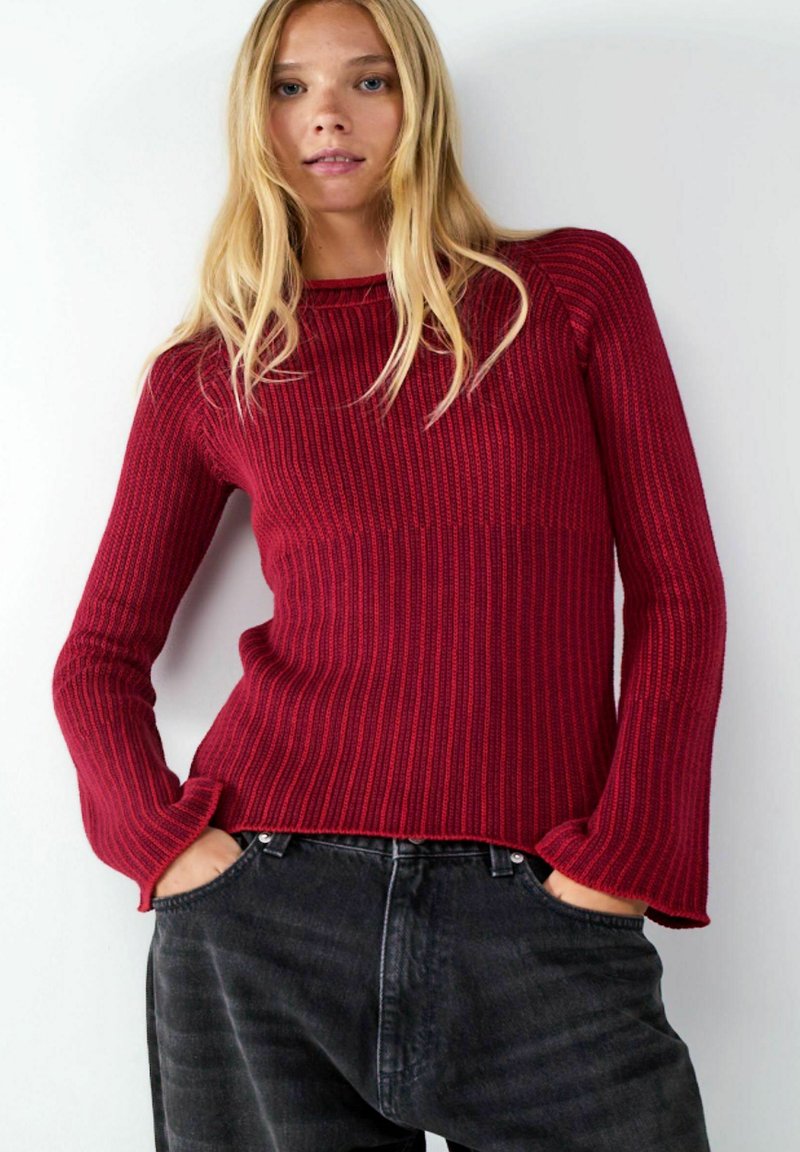 Roter gerippter Strickpullover mit rundem Halsausschnitt und langen Ärmeln, kombiniert mit hochtaillierten schwarzen Jeans in lockerer Passform.