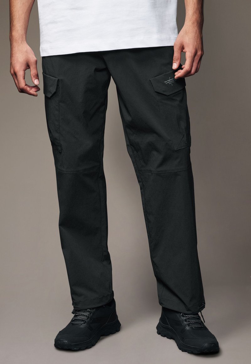 Schwarze Cargo-Hosen mit glatter Textur, die mehrere Taschen und eine lockere Passform bieten. Kombiniert mit schwarzen Wanderschuhen.