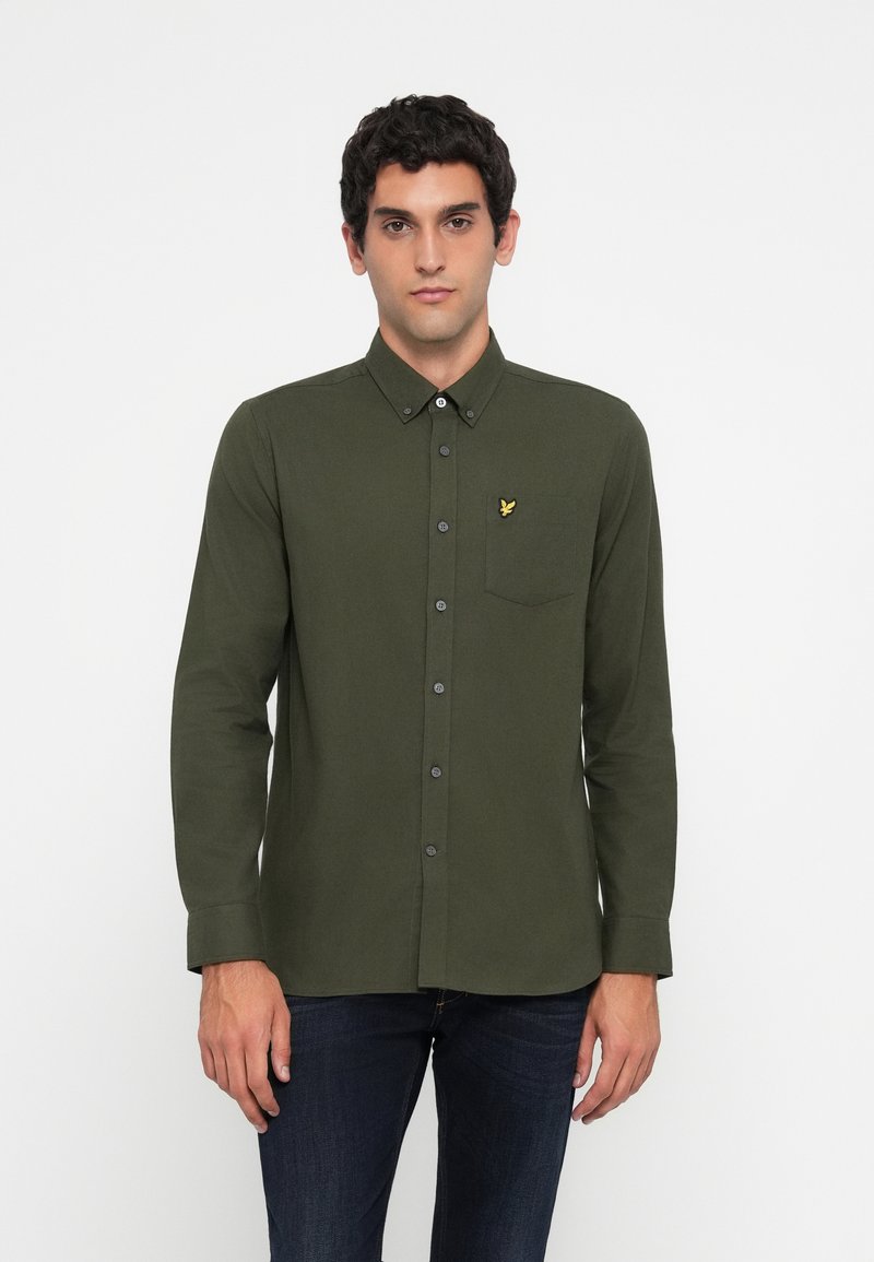 Hombre vistiendo una camisa de botones verde oscuro de manga larga con un pequeño logo de un pájaro amarillo en el bolsillo izquierdo del pecho.