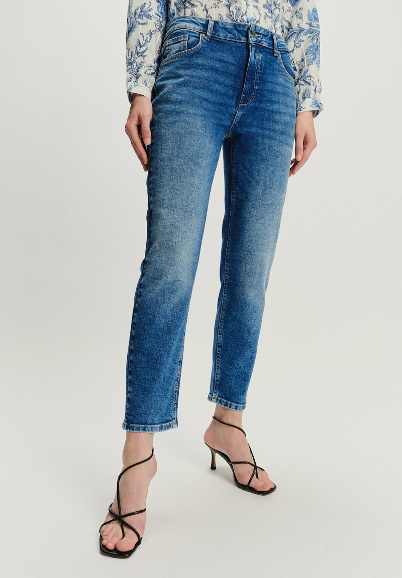 Kobieta ubrana w niebieskie, dopasowane jeansy, w połączeniu z czarnymi, wiązanymi sandałami na wysokim obcasie oraz kwiatową bluzką z długim rękawem.