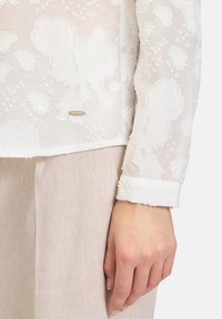 White floral-embroidered sheer blouse with long sleeves and a cuff. Features a small metallic label at the hem. Paired with beige textured pants.