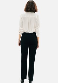 Blouse blanche à manches longues avec des poignets retroussés, associée à un pantalon noir ajusté, présentant une texture lisse et une coupe droite.