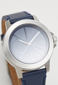 Montre argentée avec un cadran dégradé bleu et desIndex en argent. Bracelet en cuir bleu lisse avec des coutures contrastantes. Design minimaliste.