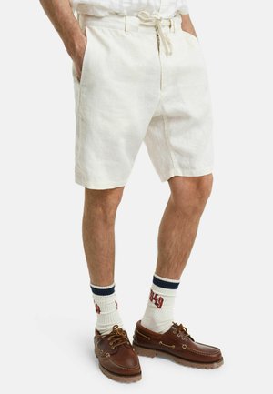 Shorts bianchi in lino presentano una vestibilità comoda, tasche laterali e una vita regolabile con cordoncino. Indossati con scarpe da barca marroni e calze a righe.