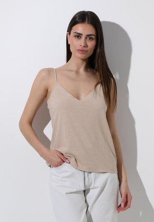 Donna con capelli castani lunghi che indossa una maglia beige senza maniche con scollo a V e pantaloni bianchi, in piedi contro un muro bianco con una mano in tasca.