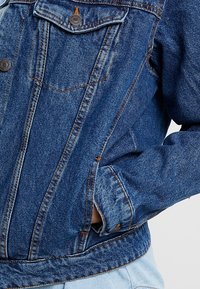 Chaqueta de denim en azul oscuro con una textura ligeramente desgastada, que presenta dos bolsillos en el pecho, botones a presión y una cintura entallada.