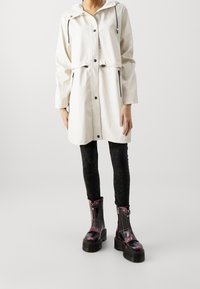 Hvid vandtæt jakke med hætte, forlommer og sorte lynlåsdetaljer, kombineret med sorte teksturerede leggings og skinnende, mønstrede ankelstøvler.