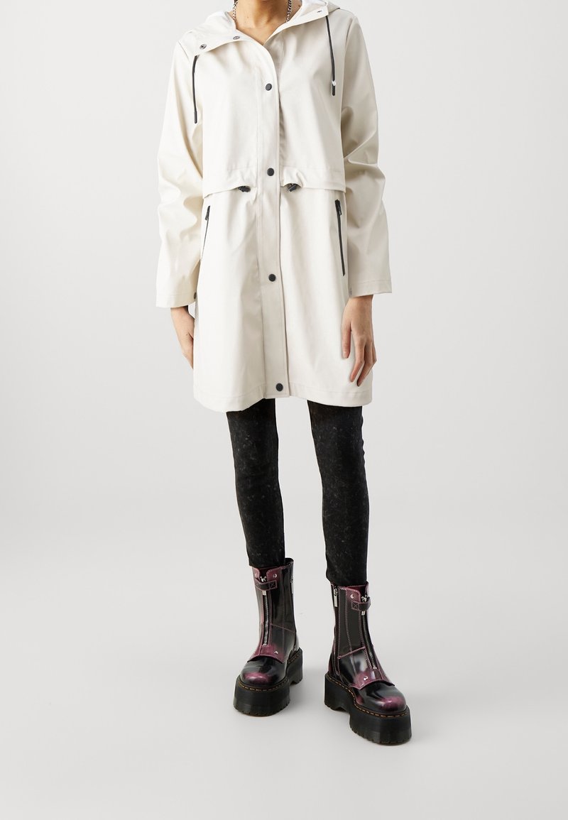 Hvid vandtæt jakke med hætte, forlommer og sorte lynlåsdetaljer, kombineret med sorte teksturerede leggings og skinnende, mønstrede ankelstøvler.