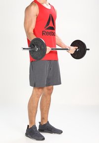 Débardeur sans manches rouge avec le logo Reebok noir, shorts gris et chaussures de sport noires, tenant une barre avec des disques de poids noirs.