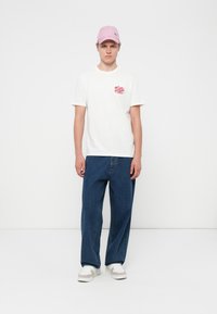 White graphic t-shirt with red logo, paired with blue loose-fit jeans and light pink cap, completed with white and beige sneakers.