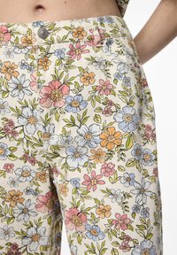 Pantalones de estampado floral con fondo beige, que presentan flores multicolores en rosa, azul y amarillo. Incluyen bolsillos delanteros y cierre de botón.