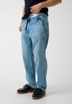 Mann trägt hellblaue Denim-Jogginghosen, schwarzes T-Shirt, schwarze Leder-Schnürschuhe und zeigt die weiße, mit Markenlogo versehene Bund seines Unterwäsche.