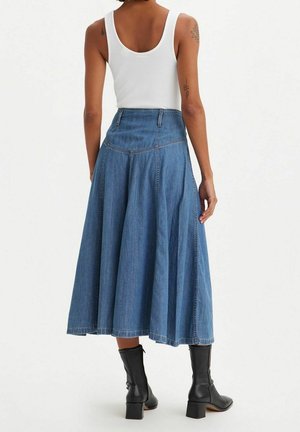 Kvinde iført en hvid ærmeløs top, blå denim midi nederdel med høj talje og sorte ankelstøvler med hæl, stående mod en ensfarvet baggrund.