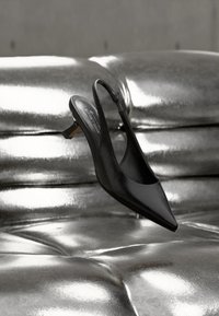 Μαύρο δερμάτινο παπούτσι με λουράκι στη φτέρνα, μυτερή μύτη και μικρό τακούνι, εκτεθειμένο σε γυαλιστερή, ασημένια επιφάνεια με σκαλίσματα. Ομαλή υφή, λιτός σχεδιασμός.