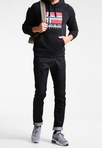 Černá mikina s kapucí s grafickým logem, doplněná tmavými džínami. Oblečení zahrnuje ohrnuté manžety a šedé tenisky. Na jednom rameni je přehozený batoh.