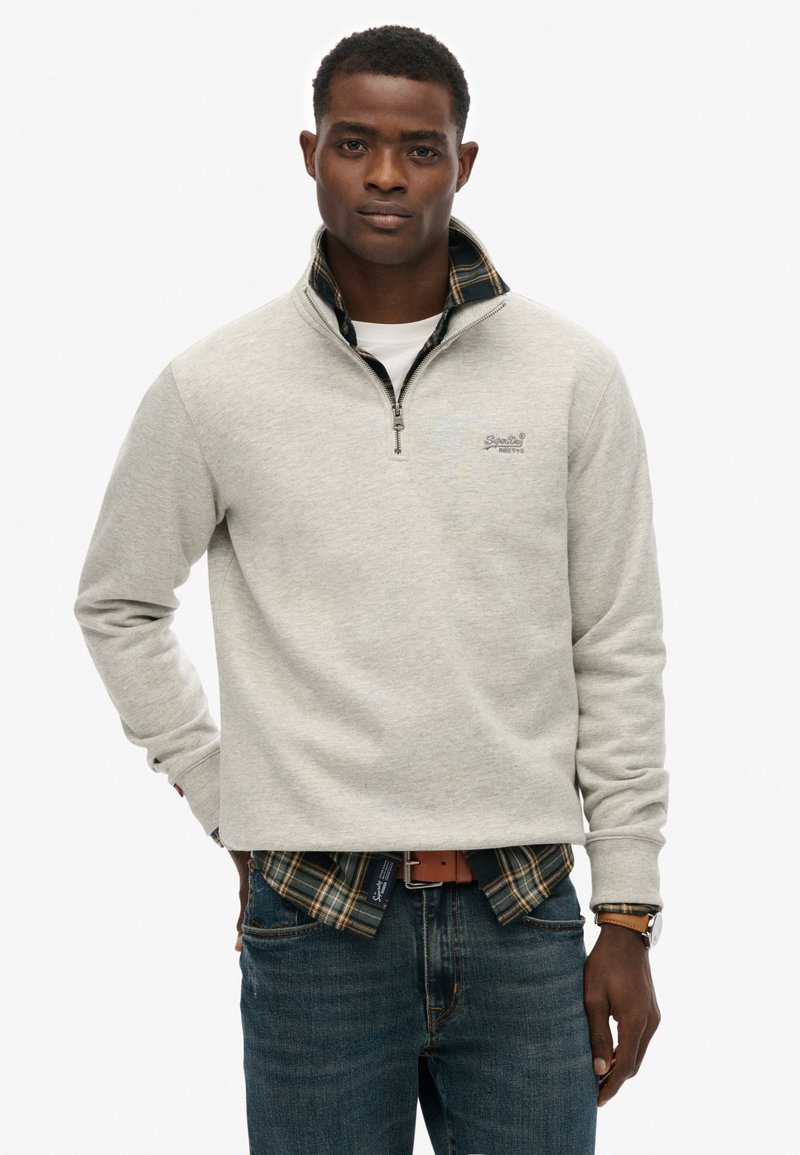 Szara bluza na zamek z wysokim kołnierzem, założona na koszulę w kratę z flaneli. Teksturowana tkanina z logo na piersi.