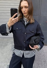 Chaqueta corta azul marino con botones plateados y dos bolsillos en el pecho, sobre una camisa de rayas en blanco y negro; sosteniendo un bolso de cuentas negras.