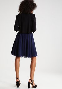 Mujer con cabello rizado, vestida con una blusa de manga larga negra, una falda azul marino a la altura de la rodilla y sandalias negras de tacón alto, de espaldas.