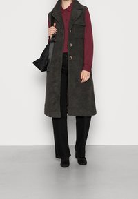 Donkergrijze, fluffy vest met een kraaglijn, knoopsluitingen en voorzakken, gecombineerd met een wijnrode blouse en zwarte broeken.