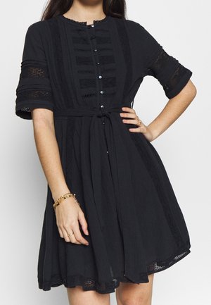 Kobieta w czarnej sukience do kolan z krótkimi koronkowymi rękawami, zapinanej na guziki z paskiem w talii oraz złotą bransoletką i pierścionkami.