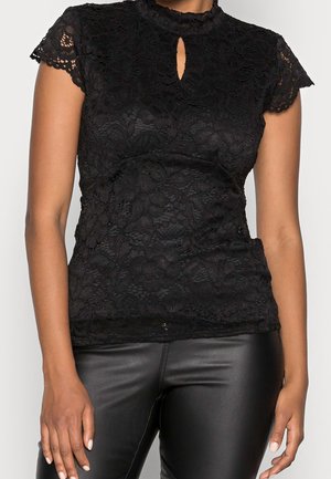 Mujer con una blusa de encaje negra de mangas cortas y escote con ojal, combinada con pantalones de cuero negro, de pie frente a un fondo liso.