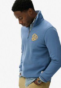 Sudadera de forro polar azul claro con media cremallera, adornada con un logo bordado en amarillo. Lleva encima una camisa de cuadros de botones con rayas verdes y blancas.