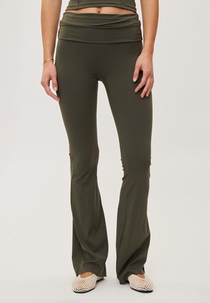 Persona che indossa leggings a zampa vita alta verde oliva e ballerine intrecciate color crema, in piedi su uno sfondo chiaro e semplice. È visibile solo la parte inferiore del busto.