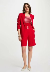 Blazer rosso su misura sopra una maglietta a righe rossa e bianche, abbinato a pantaloni corti rossi. Presenta una cintura con fibbia argentata e scarpe beige.