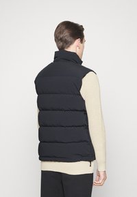 Černá prošívaná vesta s vysokým límcem a horizontálním prošíváním. Nositelná přes krémový pletený svetr, s batohem na zpět.